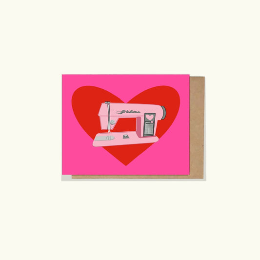 A greeting card with a brightly colored pink background and a vintage ight pink sewing machine on the background of a red heart. 