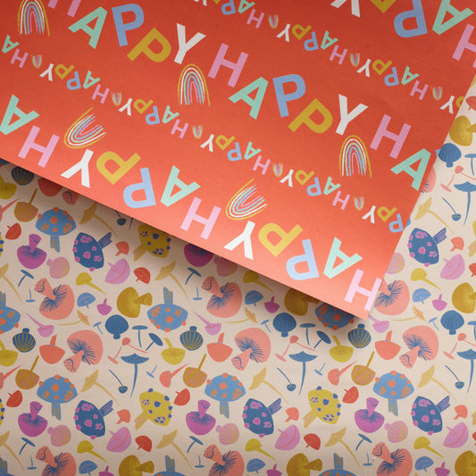 Double-sided gift wrapping paper with a pattern of brightly colored mushrooms on one side and the word 'HAPPY' written in a colorful font on the reverse, placed on a flat surface.

